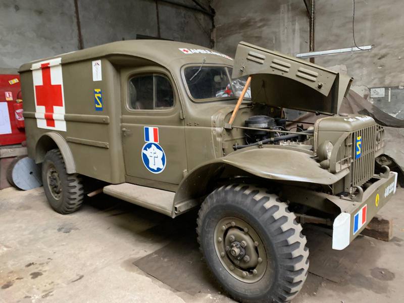 L'ambulance Dodge restaurée dans un hangar, capot ouvert, croix rouge et cocarde de la 2e DB sur la portière.
